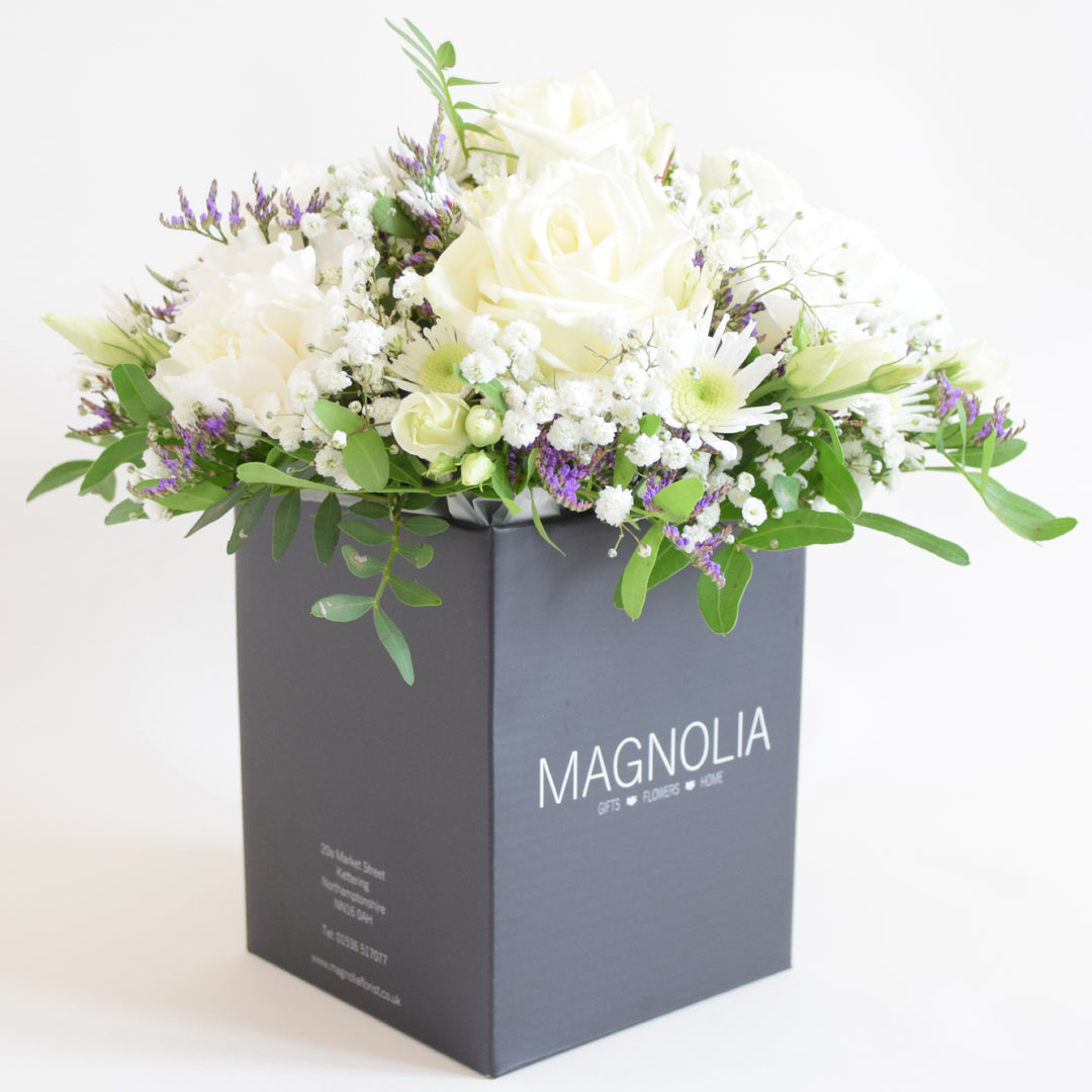 Dainty Kew Arrangement - Magnolia the Florist The Kew Arrangement from Magnolia the Florist, features a stunning mix of white florals with a hint of purple, including roses, carnations, lisianthus, gyp, chrysanthemum and limonium, all arranged in floral foam. Presented in a signature box, it makes a beautiful and sophisticated gift for any occasion. For scale purposes, the box container measures approximately 15cm x 12cm x 12cm, giving the overall arrangement size of 25cm h x 18cm w approximately.