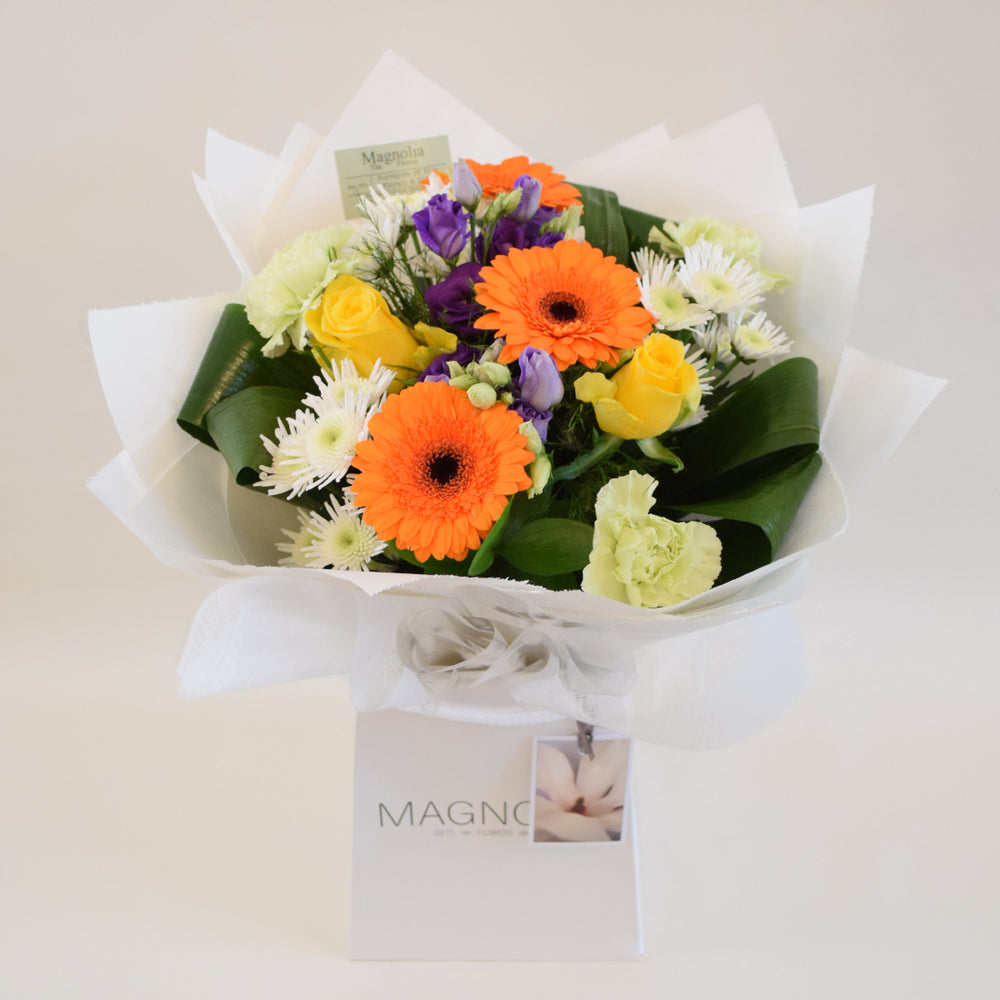 Savoy Hand-Tied Bouquet - Magnolia the Florist With vibrant hues of yellow, orange, green, and purple, the Savoy Hand-Tied Bouquet from Magnolia the Florist, is a stunning mix of Roses, Mini Gerbera, Lisianthus, Carnations, Chrysanthemums, and foliage. Expertly arranged in our signature box bags, this aqua bouquet is sure to brighten any space. Perfect for any occasion, bring a touch of nature's beauty into your home today.